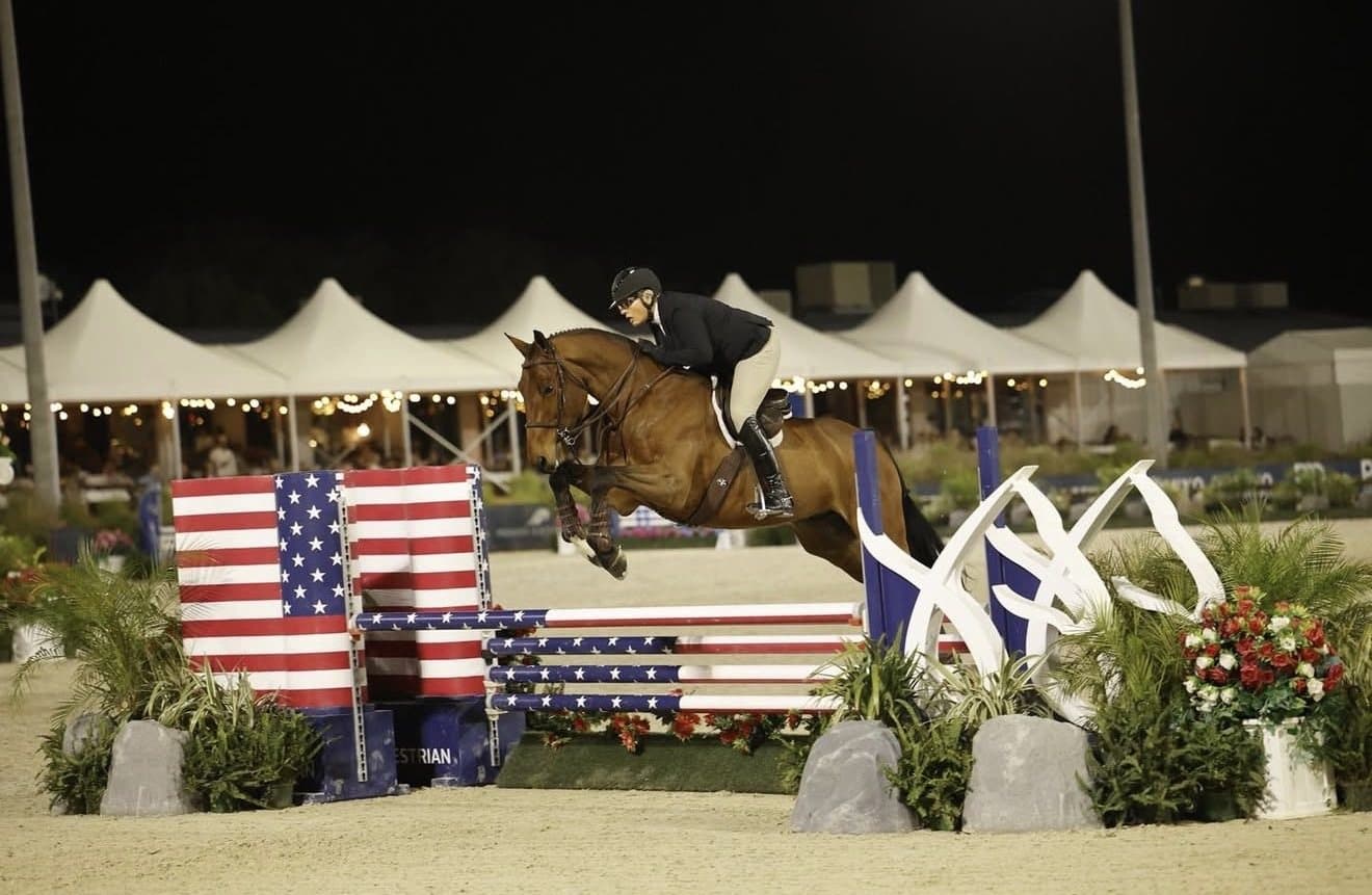 A horse and rider jumping under the lights at an evening show