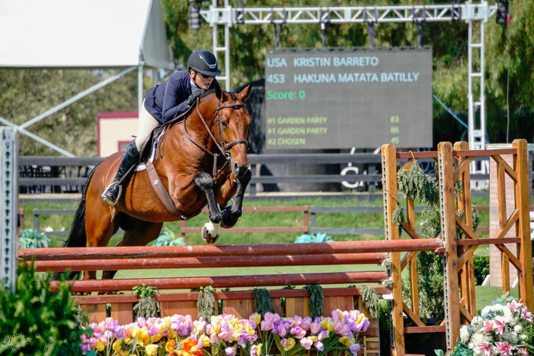 Kristin Barreto riding Hakuna Matata Batilly over a fence, her name on the scoreboard behind
